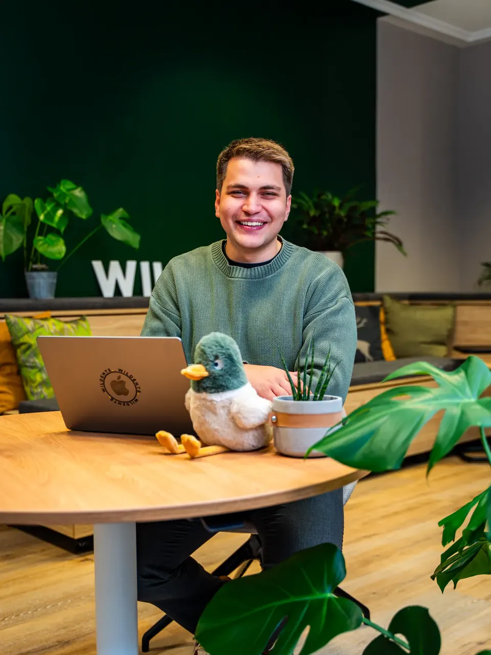 Teammitglied lacht am Laptop im Wildente-Büro, Plüschente auf dem Tisch vor grüner Wand