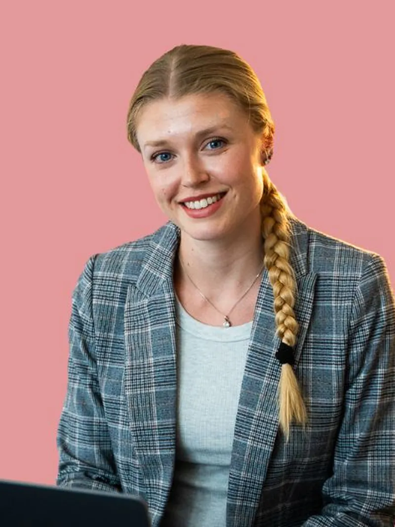 Alex mit geflochtenem Zopf lächelt am Laptop, rosafarbener Hintergrund