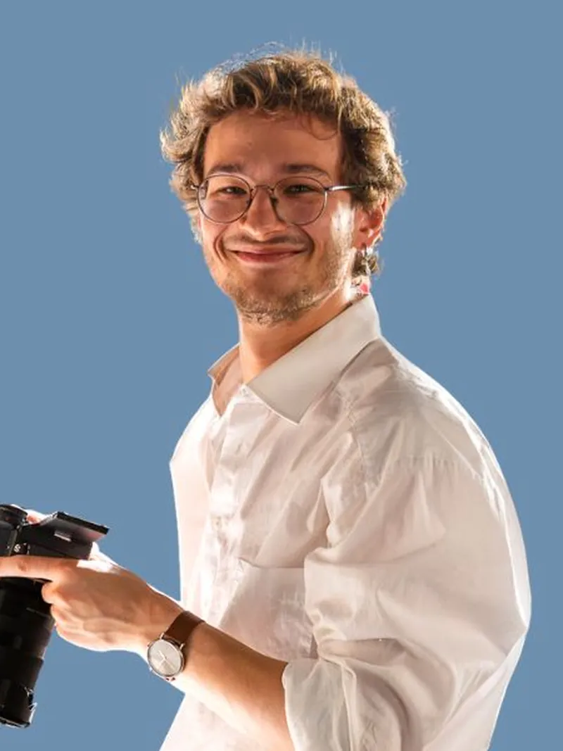 Maksim mit Brille und Kamera in der Hand vor blauem Hintergrund